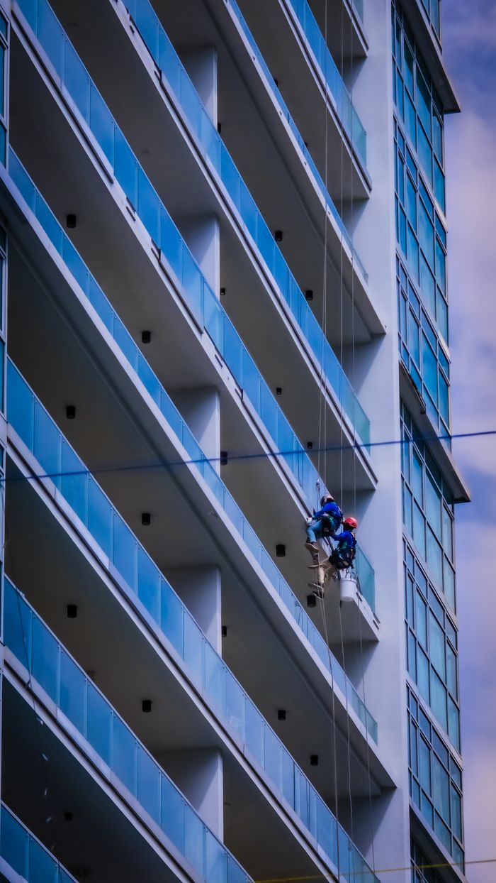 Two window cleaners suspended on ropes working on a high-rise building facade under a sunny sky.